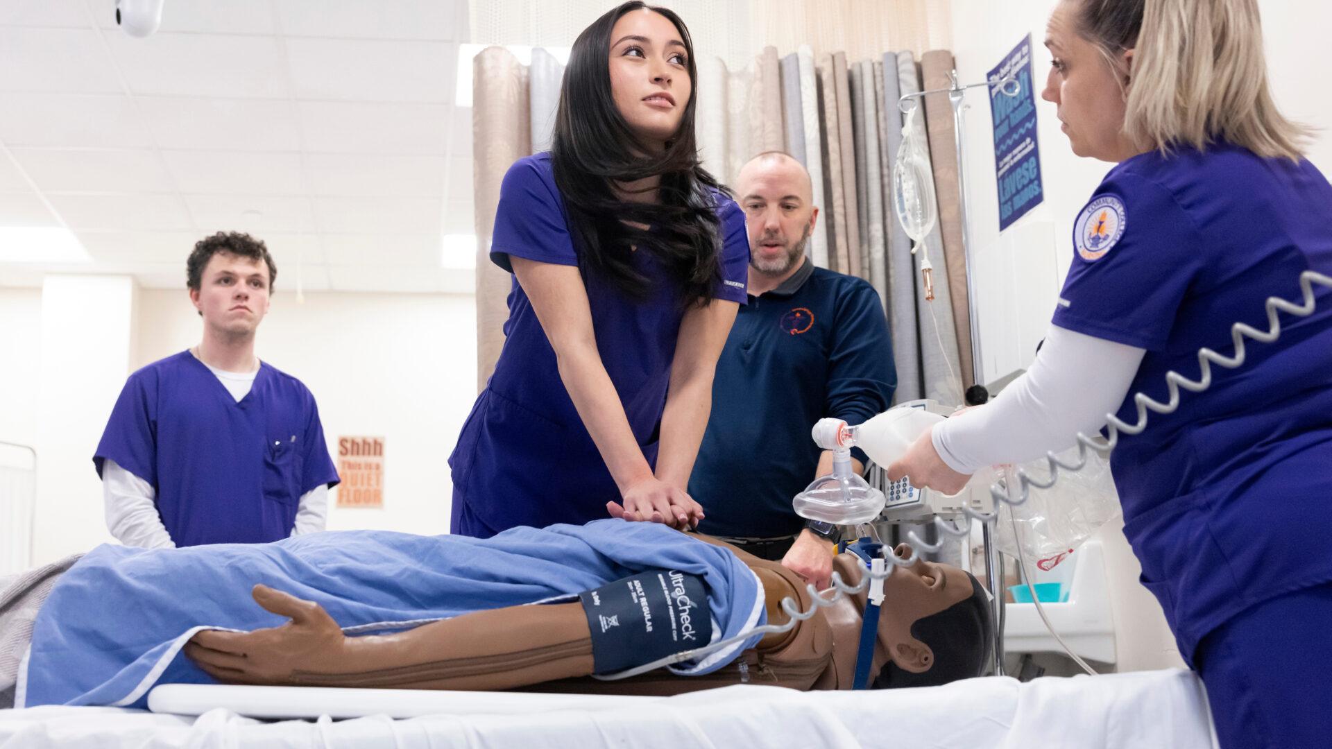 Female nursing student giving chest compressions during a CPR lesson in a lab classroom on a college campus.