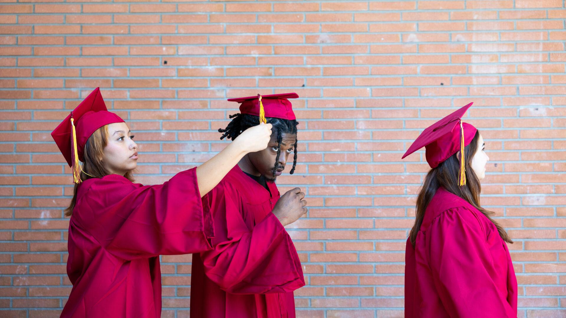 Two Hispanic female students in graduation cap and gown adjusting the mortarboard of a Black male student in preparation for a graduation ceremony on a college campus.